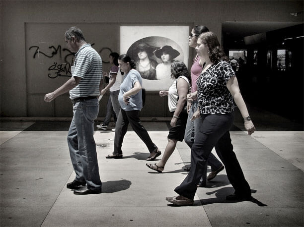 Foto do mesmo local enquadrado de frente, porém mais de longe. A foto antiga está ao fundo. No centro da imagem estão passando seis pessoas da direita para a esquerda, de perfil, andando separadas, em diferentes distâncias. Elas olham para a frente ou para baixo, exceto por uma mulher que parece olhar a foto antiga. As duas mulheres na foto antiga estão visíveis, bem no meio desse grupo de pessoas passando.