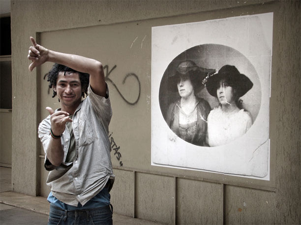 Foto do mesmo local enquadrado de frente, de perto, com a parede em perspectiva. Na metade direita está a foto antiga. Na metade esquerda, em primeiro plano, há um rapaz olhando para a câmera e posando para a foto, fazendo um hang loose com cada mão e dando um pequeno sorriso. Uma das mãos está acima da cabeça, com braço e antebraço fazendo um ângulo reto. A outra mãoestá na frente do peito. Ele usa dreads curtos; sua postura é graciosa e seu olhar é um pouco inseguro. O rapaz aparece do alto das coxas para cima e seu corpo encobre parcialmente algumas pixações na parede.