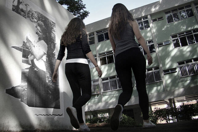 Foto horizontal do mesmo local, com o mesmo enquadramento rente ao chão. As mesmas adolescentes, com a mesma roupa, estão passando de volta, de costas, em primeiro plano, no centro da foto. À esquerda delas a foto antiga aparece rasgada pela metade, de cima a baixo, e não se vê mais o bebê.