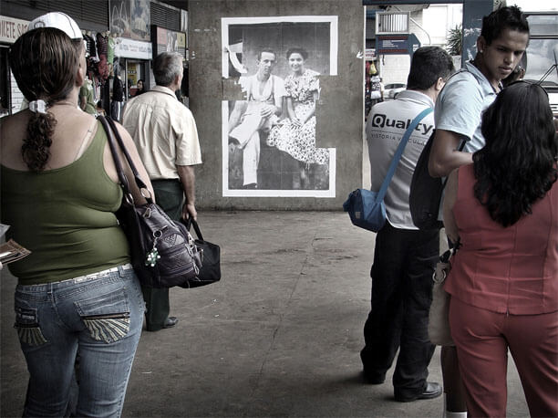 Foto horizontal mostrando seis pessoas, três à esquerda e três à direita, como se formassem duas filas paralelas. No centro, ao fundo, está colada uma foto antiga numa larga coluna de concreto. O lugar parece ser uma rodoviária; vê-se parte de um ônibus na margem direita e parte de uma fileira de lojinhas na margem esquerda. As três pessoas da "fila" à esquerda estão olhando a foto antiga; o homem da frente, de meia idade, grisalho, se inclina para a frente, parecendo curioso.A foto antiga é vertical e mostra um jovem casal sentado, olhando para a câmera. A mulher sorri, com as mãos sobre o colo; ela usa um vestido claro com pequenas estampas. O homem usa camisa de mangas curtas e suspensório. Eles estão ligeiramente inclinados um para o outro. Pequens faixas da foto foram arrancadas nas margens laterais.