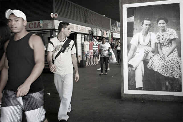 Foto horizontal; a foto antiga aparece maior, à direita, junto à margem. Ao lado e ao fundo há algumas pessoas e as lojinhas. Um homem de boné está passando no lado esquerdo, já quase saindo da margem, de frente para a câmera e bem perto. Ele olha para a câmera com uma expressão meio agressiva, meio de desdém. Atrás dele há outro homem, também andando em direção à câmera, mas com a cabeça virada para olhar a foto, que está ligeiramente atrás dele.