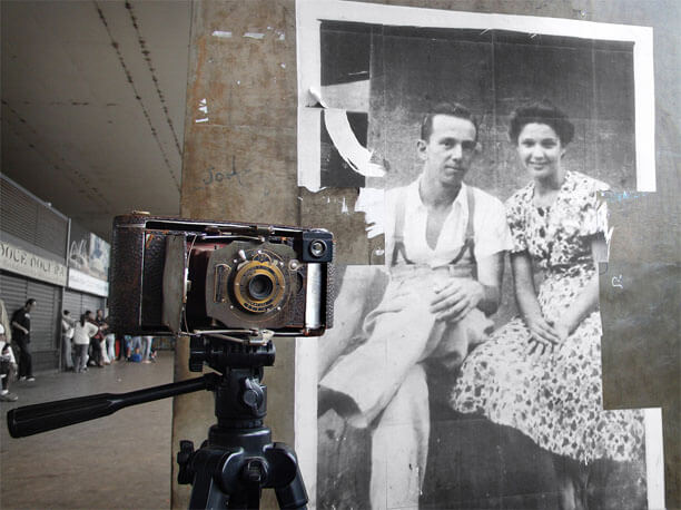 Foto horizontal mostrando em quase todo o quadro a coluna de concreto com a foto antiga. À esquerda vê-se uma estreita faixa do fundo em perspectiva, com pessoas ao longe. Em primeiro plano há uma câmera fotográfica muito velha, que aparenta ser do começo do século passado. Ela aparece de frente, a meia altura, apoiada num tripé, com um dos lados se sobrepondo à foto antiga.