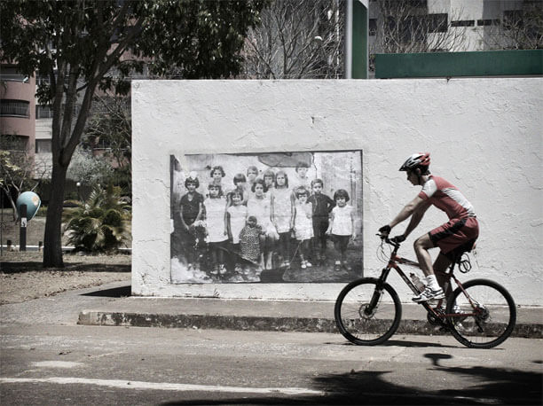 Foto horizontal de um homem passando de bicicleta, de lado para a câmera, da direita para a esquerda. Ele usa roupas de ciclista e um capacete de formato futurista, próprio para esse esporte. Suas roupas, capacete e bicicleta são vermelhas com detalhes brancos. Está em frente à parede onde está colada uma foto antiga (a mesma das imagens anteriores). A roda da frente da bicicleta está junto ao canto da foto antiga. Atrás da parede vê-se uma árvore, um orelhão, algumas plantas e dois prédios.