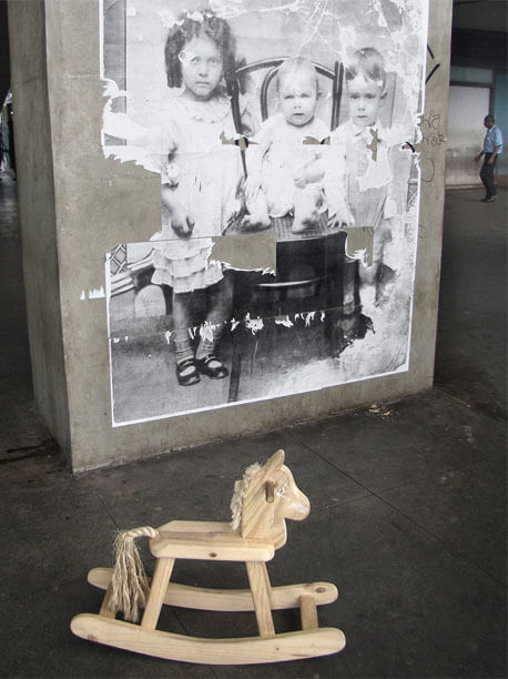 Foto vertical mostrando a parte de baixo da coluna com a foto antiga colada. Parte da margem esquerda foi arrancada, assim como pequenos pedaços no centro da imagem, mas ainda pode-se ver bem as crianças. À direita da coluna aparece um pedacinho do fundo, onde se vê bem ao longe um homem de perfil, entrando no quadro. Em primeiro plano, no chão escuro em frente à coluna, está um cavalinho de pau de madeira clara, desses em que crianças pequenas se sentam e balançam.