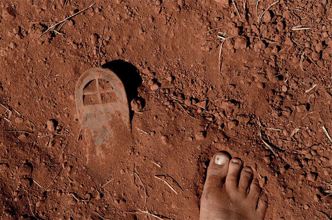 Foto horizontal mostrando a sombra de uma mão indo em direção à saída de um cano de PVC, com cerca de 10cm de diâmetro. O cano está num chão de terra marrom-rosado, com manchas brancas de líquido seco. Ele está perpendicular à margem de cima da foto, à esquerda. A sombra do braço sai do canto direito e faz uma diagonal até quase a boca do cano; um dos dedos "toca" uma sombra que faz uma faixa preta em toda a extensão superior da imagem. O sol que entra pela esquerda ilumina o chão e a saída do cano, cujo interior também está preto. 
