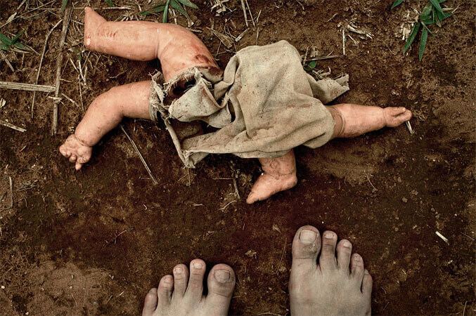 Foto horizontal; detalhe de um chão marrom escuro com alguns pedaços de capim seco. Há um corpo sem cabeça, sujo de terra, de algo que já foi uma boneca mas agora é sinistro: uma perna e um braço de plástico à esquerda, um tecido embolado, rasgado e sem recheio no meio, um braço e um pé à direita. Está na metade da imagem, de perto, na parte de cima. Na margem de baixo está a metade de dois pés descalços e muito sujos.