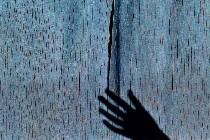 Foto horizontal de uma superfície de madeira pintada de azul turquesa, coberta de finas rachaduras no sentido vertical. No centro, uma dessas rachaduras é maior, uma abertura preta com um centímetro de largura, que se afina nas pontas. A sombra de uma mão sai em diagonal da margem de baixo da foto, à direita. Quatro dedos da sombra tocam a parte de baixo da abertura.
