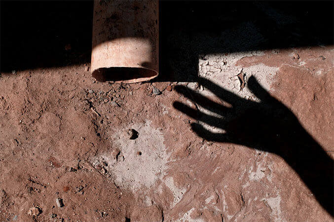 Foto horizontal mostrando o detalhe de um chão de terra vermelha e seca, com alguns torrões e pedacinhos de capim seco. No lado direito, embaixo, aparece metade de um pé descalço, afundando na terra solta. No lado esquerdo aparece a sola de um sapato, parciamente coberta de terra.