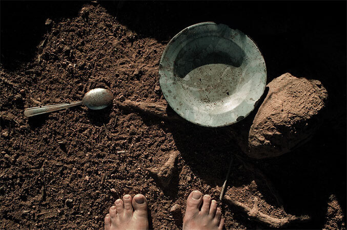 Foto horizontal; detalhe de um chão de terra marrom onde metade de dois pés descalços surgem na margem de baixo, defronte a um prato de ferro velho e enferrujado, com uma colher de sopa ao lado. Aparecem partes da raiz de uma árvore no chão. A cena está iluminada pelo sol do final do dia, com sombras pretas e alongadas em grande parte da  imagem, na direção da margem de cima para a de baixo. 