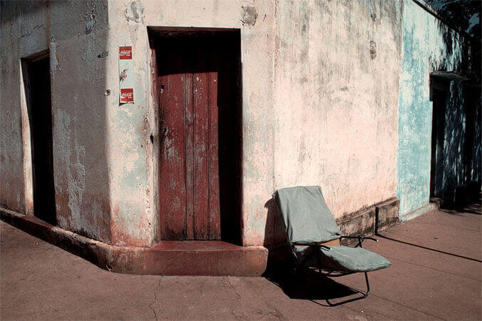 Foto horizontal de uma espreguiçadeira velha, vazia, junto a uma fachada com uma porta fechada. A calçada é marrom, de cimento; a parede é azul no canto direito e branca no restante, com pintura velha e muito manchada. A fachada fica numa esquina e a porta está numa estreita parte chanfrada; é de madeira, pintada de marrom avermelhado e muito velha. Nas paredes dos dois lados vê-se outras duas portas (apenas um vão feito pela sombra do sol a pino). A espreguiçadeira também faz uma sombra preta no chão, e tem almofadas encapadas de tecido azul acinzentado. Não há ninguém na rua.