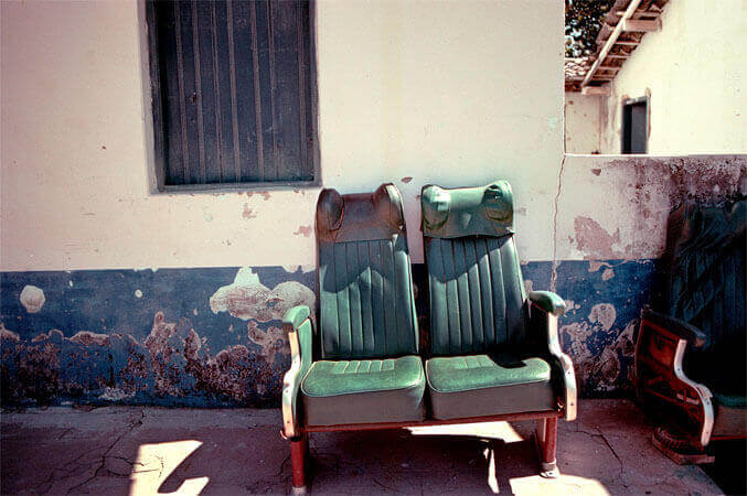 Foto horizontal de duas poltronas de ônibus, velhas e vazias, com estofamento de couro verde. Elas estão sobre uma calçada de cimento rachado, encostadas na fachada de uma casinha. A casinha tem parede branca com uma faixa azul escura embaixo, que está descascada e manchada. Acima das poltronas, à esquerda, aparece uma janela fechada, estreita e de madeira escura, cortada na margem de cima. Do lado delas há parte de outra poltrona, cortada na margem direita.