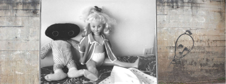 Colagem digital em formato panorâmico. No centro há uma foto horizontal preto e branca de duas bonecas sobre uma colcha, encostadas a uma parede. Uma delas é de pano, com corpo de tecido branco com bolinhas escuras; a cabeça é oval, preta e sem cabelo, com grandes olhos redondos. A outra é de plástico, com pés amassados e cabelos louros. As duas bonecas estão muito usadas; cada uma parece vestir uma peça de roupa velha de um bebê. Do lado direito há uma foto quadrada de uma parede de concreto marrom com manchas escuras, onde está grafitada uma cabeça de forma curiosa, como a de um boneco de meia, triste e cabisbaixo. Do lado esquerdo da colagem há uma estreita faixa vertical mostrando um detalhe da parede de concreto.