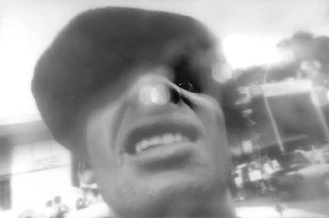 Foto horizontal do rosto de um homem de boné, do queixo para cima, ocupando quase todo o quadro. Ele olha para a câmera de cima, com uma expressão de real agressividade, mostrando os dentes, com a boca aberta e curvada para baixo. Do nariz para cima o rosto está escuro pela sombra do boné, exceto pelo olho direito, arregalado e fixo na câmera. Ao fundo, longe, vê-se pessoas e um prédio. Um anteparo na lente cria algumas formas brancas, pequenas e arredondadas, duas delas sobre o nariz do homem. O anteparo deixa tudo desfocado, exceto por um pequeno buraco onde se vê o olho nitidamente.