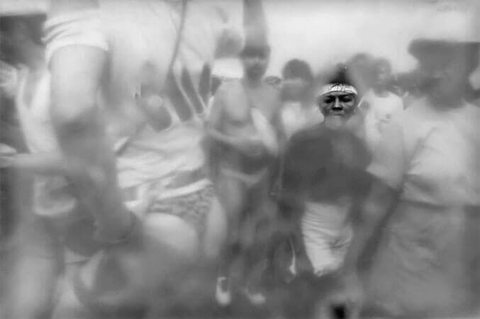 Foto horizontal de várias pessoas num bloco de carnaval. Na metade da direita há um grupo passando, de frente para câmera. Na metade da esquerda há um homem de costas, sambando com uma mão no quadril e a outra levantada. Ele está bem perto, cortado do ombro para cima e da coxa para baixo. Usa uma camiseta curta e uma tanga com estampa de onçinha que deixa a bunda quase toda de fora. No grupo que está passando ao lado dele há duas mulheres, mais próximas à câmera, que olham para a sua bunda. O anteparo na lente deixa tudo desfocado, menos um pequeno buraco onde se vê nitidamente o rosto da segunda mulher, que está séria.