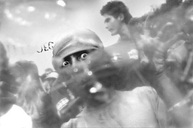 Foto horizontal de um bloco vindo em direção à câmera, com enquadramento inclinado para a esquerda. Há um rapaz em primeiro plano, com um pano amarrado na cabeça, logo acima dos olhos. Ele faz uma pose cômica, com os olhos muito arregalados, os lábios em bico e as duas mãos em garra. Está bem perto e aparece do peito para cima. O anteparo na lente deixa tudo desfocado, menos uma pequena faixa onde se vê os olhos arregalados.