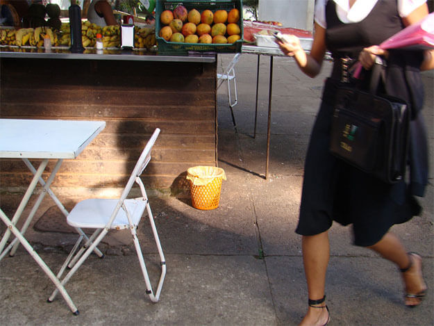 Foto horizontal de uma mulher passando apressada na rua, cortada dos ombros para cima. Ela usa vestido preto sobre uma blusa branca de mangas curtas, calça sandálias pretas e o pé que está na frente está cortado pela margem de baixo da foto. Está entrando na imagem pela margem direita, e carrega várias coisas nas duas mãos (celular, bolsa e casaco pretos, duas pastas rosas). O chão é um pátio de cimento. Na metade esquerda da foto há um quiosque de frutas, visto do chão até o balcão, onde estão expostos mamões e bananas. Na frente do quiosque aparece metade de uma mesinha branca de metal com uma cadeira. Bem no centro da imagem há um cesto de lixo laranja vivo, iluminado pela luz do sol do começo da manhã. A luz bate em outras pequenas partes da foto.