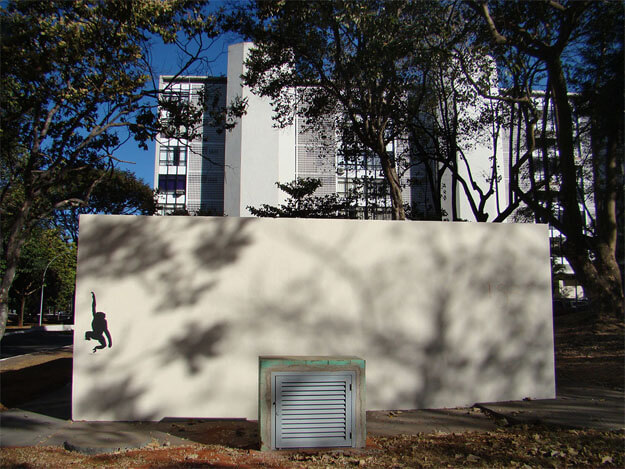 Foto horizontal mostrando, no centro, uma parede bege, retangular, com cerca de 6m de largura por 2,5m de altura. Ela ocupa quase todo o espaço na horizontal, e metade do espaço na vertical. Está iluminada pelo sol, com sombras de árvores fazendo desenhos, e junto ao seu lado esquerdo foi pintada a silhueta de um macaco pequeno, preto, pendurado por um braço. Diante da parede há uma calçada, um cubo de cimento de 1m por 1m com uma portinhola cinza e uma pequena faixa de chão de terra com folhas secas. Ao fundo vê-se grandes árvores, o céu azul vivo e um prédio branco, baixo e comprido, iluminado pelo sol.