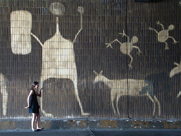 Foto horizontal mostrando o detalhe de um paredão com cerca de 5m de altura, revestido de pequenos ladrilhos compridos, assentados na vertical. O paredão está marrom escuro de sujeira, quase preto. Nele há grandes desenhos brancos feitos com a técnica de arte reversa (limpando em vez de pintar). Os desenhos imitam arte rupestre, com imagens de animais e uma enorme figura humana que parece segurar um escudo. Há uma jovem de vestido preto curto e sapatilhas pretas no canto da esquerda, junto à margem de baixo. Ela está de perfil, sorrindo e fazendo uma pose que imita os gestos de pessoas em pinturas do Egito Antigo.