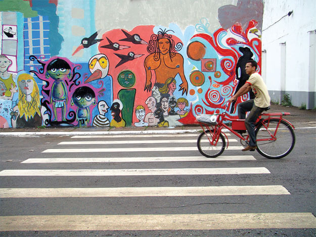 Foto horizontal de uma rua de asfalto com uma faixa de pedreste branca enquadrada de frente. Um homem está passando de bicicleta, da direita para a esquerda. Do outro lado da rua há uma calçada e junto a ela uma parede coberta por pinturas coloridas de vários artistas, em cores vivas, mostrando vinte pessoas bem diferentes umas das outras, alguns pássaros e um fundo com cores e formas diversas.