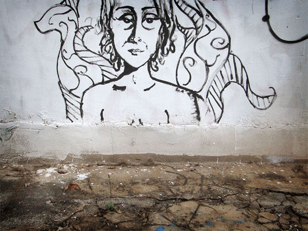 Foto horizontal mostrando o detalhe de uma parede branca, velha e manchada, e um chão de cimento escuro, velho e rachado. Na parede, bem no centro da imagem, está uma pintura a pincel com tinta preta. Ela mostra uma pessoa séria, de olhar triste, dos ombros para cima, olhando para a câmera. Está cortada da testa para cima. Atrás da pessoa há diversas formas curvas que lembram tentáculos ou asas.