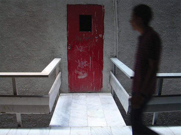 Foto horizontal da silhueta de uma pessoa passando de perfil, borrada, do lado direito, dos joelhos para cima. No centro há uma porta vermelha
                vista de frente, a cerca de três metros de distância. A porta tem a pintura de cor viva mas manchada com respingos brancos e algumas pixações. A parede é de cimento chapiscado. Tudo está na sombra, menos o chão de mármore branco e o guarda-corpo de uma estreita passarela que vai do chão até a porta.