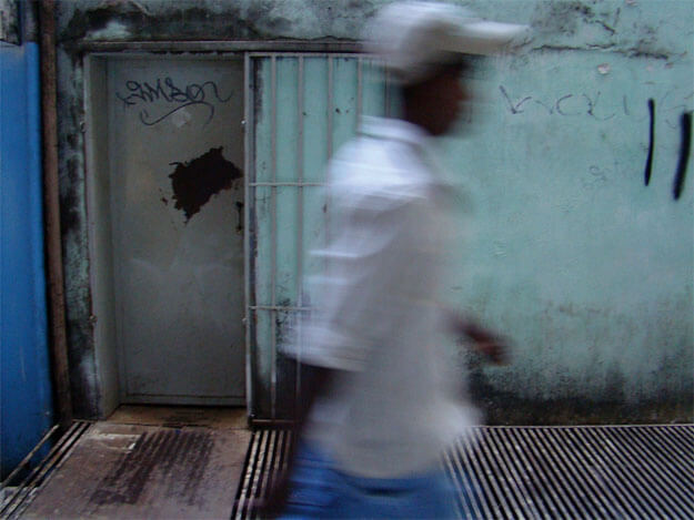 Foto horizontal de um homem passando de perfil, borrado, no centro da imagem, do quadril para cima. Ele usa calça azul, camisa branca e boné cinza clarinho. Atrás dele há uma porta cinza com uma grande mancha preta de ferrugem e um nome pixado. Ela está numa parede verde clara com pintura muito velha, manchas pretas de umidade e algumas pixações. Numa estreita faixa à esquerda a parede está pintada de azul novo e vivo, no mesmo tom da calça do homem. No chão há uma grade de ventilação enferrujada, acompanhando a linha da parede.