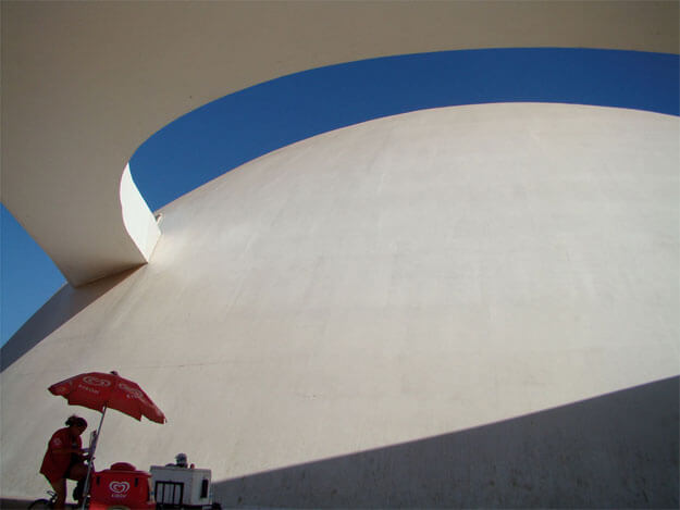 Foto horizontal do Museu da República, mostrando parte de seu formato em semicírculo e parte de uma rampa curva no alto. A rampa sai do lado esquerdo do prédio atravessa toda a imagem na horizontal. Entre o museu e a rampa vê-se uma estreita faixa céu azul vivo, sem nuvens. No canto esquerdo, embaixo, há uma vendedora de picolé num carrinho vermelho, vestida de vermelho, com um guarda-sol vermelho. Ela está a meia distância, na sombra. A sombra começa no canto esquerdo e se abre numa faixa triangular até a margem direita.